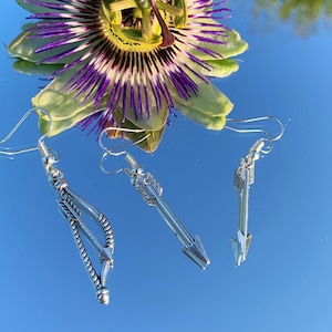 May include: Three silver arrow earrings with a feather detail. The earrings are hanging from a reflective surface.