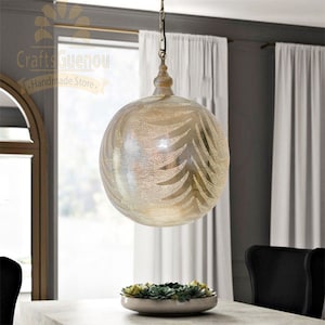 Puede incluir: Una lámpara redonda colgante de plata con un patrón decorativo recortado. La lámpara está colgada de una cadena en una habitación con cortinas blancas.