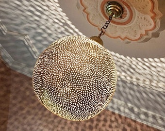 Op de afbeelding: Een ronde, decoratieve lichtarmatuur in goudkleur met een geperforeerd patroon. De armatuur hangt aan een ketting en werpt een gepatroneerde schaduw op het plafond.