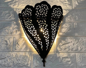 May include: A black metal wall sconce with a decorative, intricate cut-out design. The sconce is lit and casts a warm glow on the white brick wall behind it.