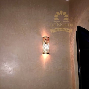 K&ouml;nnte beinhalten: Eine goldene Wandleuchte aus Metall mit geometrischem Muster und einer Gl&uuml;hbirne, die den Raum beleuchtet. Die Leuchte ist an einer beigefarbenen Wand montiert. Der Text "CraftsGuenou - Handmade Store." ist im Bild sichtbar.