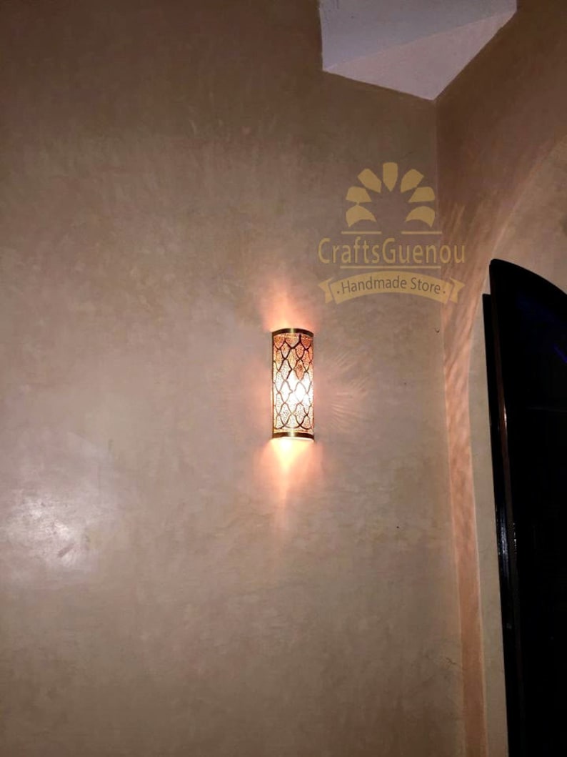 K&ouml;nnte beinhalten: Eine goldene Wandleuchte aus Metall mit geometrischem Muster und einer Gl&uuml;hbirne, die den Raum beleuchtet. Die Leuchte ist an einer beigefarbenen Wand montiert. Der Text "CraftsGuenou - Handmade Store." ist im Bild sichtbar.