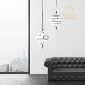 May include: Two silver Moroccan pendant lights with clear glass panels hang from chains against a white wall. A black leather sofa sits in the foreground.