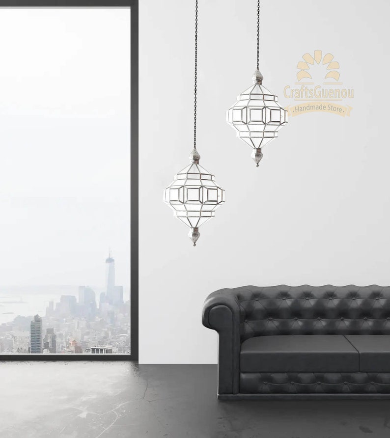 May include: Two silver Moroccan pendant lights with clear glass panels hang from chains against a white wall. A black leather sofa sits in the foreground.