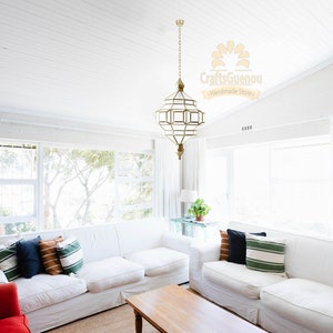 May include: A white living room with two white sofas, a wooden coffee table, and a large gold geometric pendant light hanging from the ceiling. The sofas have pillows in various colours and patterns, including green, blue, and orange. The windows are covered with white curtains. The text "Craftsguenou Handmade Store" is visible in the upper right corner of the image.