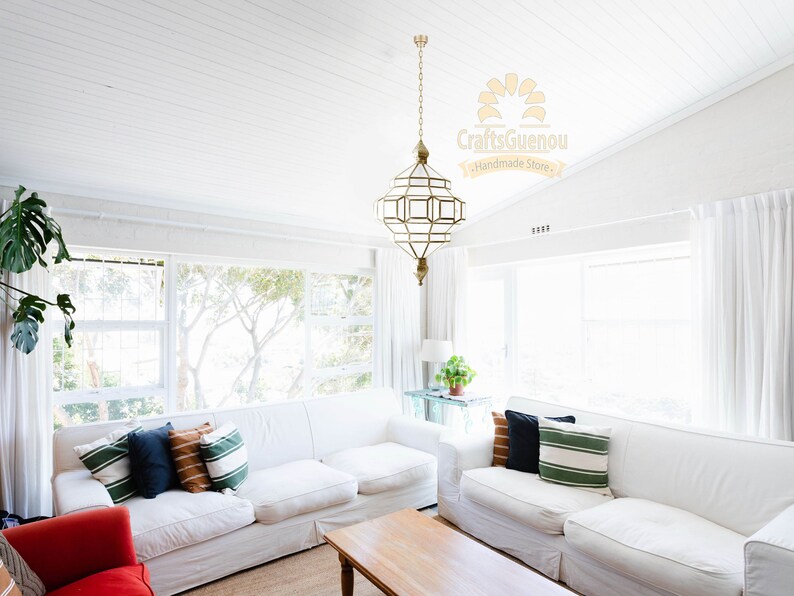 May include: A white living room with two white sofas, a wooden coffee table, and a large gold geometric pendant light hanging from the ceiling. The sofas have pillows in various colours and patterns, including green, blue, and orange. The windows are covered with white curtains. The text "Craftsguenou Handmade Store" is visible in the upper right corner of the image.