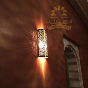K&ouml;nnte beinhalten: Eine goldene Wandleuchte aus Metall mit marokkanischem Design. Die Leuchte ist beleuchtet und wirft einen warmen Schein an die Wand. Die Leuchte ist mit "CraftsGuenou - Handmade Store - " beschriftet.
