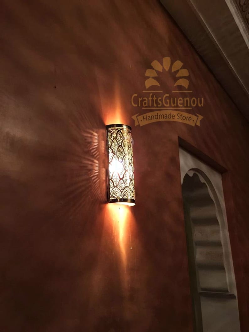 K&ouml;nnte beinhalten: Eine goldene Wandleuchte aus Metall mit marokkanischem Design. Die Leuchte ist beleuchtet und wirft einen warmen Schein an die Wand. Die Leuchte ist mit "CraftsGuenou - Handmade Store - " beschriftet.