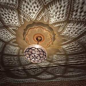 May include: A round, metal, Moroccan-style chandelier with a geometric pattern hangs from a ceiling with a decorative, white, patterned design. The chandelier casts a shadow on the ceiling.