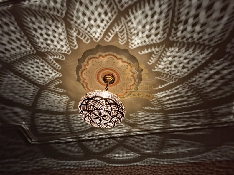 May include: A round, metal, Moroccan-style chandelier with a geometric pattern hangs from a ceiling with a decorative, white, patterned design. The chandelier casts a shadow on the ceiling.