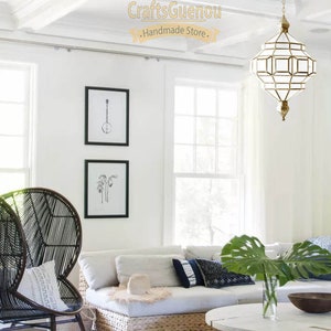 May include: A white living room with a white sofa, a black wicker chair, a white coffee table, and a large gold geometric chandelier hanging from the ceiling. The room has white walls and a white rug with a brown cowhide rug on top. The text "CraftsGuenou - Handmade Store -" is visible on the ceiling.