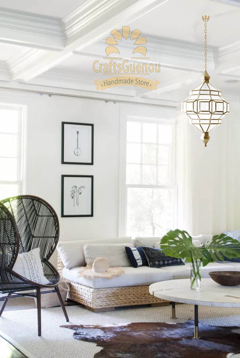 May include: A white living room with a white sofa, a black wicker chair, a white coffee table, and a large gold geometric chandelier hanging from the ceiling. The room has white walls and a white rug with a brown cowhide rug on top. The text "CraftsGuenou - Handmade Store -" is visible on the ceiling.