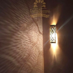 K&ouml;nnte beinhalten: Eine goldene Wandleuchte aus Metall mit geometrischem Muster und einer Gl&uuml;hbirne, die den Raum beleuchtet. Die Leuchte wirft einen dekorativen Schatten an die Wand. Der Text "CraftsGuenou Handmade Store" ist oben in der Mitte des Bildes sichtbar.