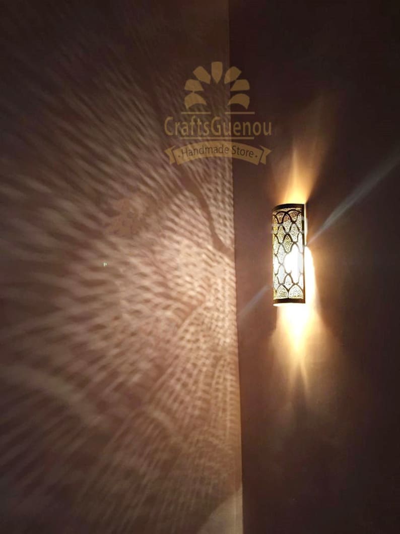 K&ouml;nnte beinhalten: Eine goldene Wandleuchte aus Metall mit geometrischem Muster und einer Gl&uuml;hbirne, die den Raum beleuchtet. Die Leuchte wirft einen dekorativen Schatten an die Wand. Der Text "CraftsGuenou Handmade Store" ist oben in der Mitte des Bildes sichtbar.