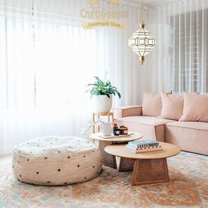 May include: A living room featuring a light pink sofa, a white fluffy ottoman, and two round wooden coffee tables. The room has a large window with white sheer curtains and a white ceiling with a gold geometric pendant light hanging from it. The floor is covered in a colourful rug with a floral pattern.