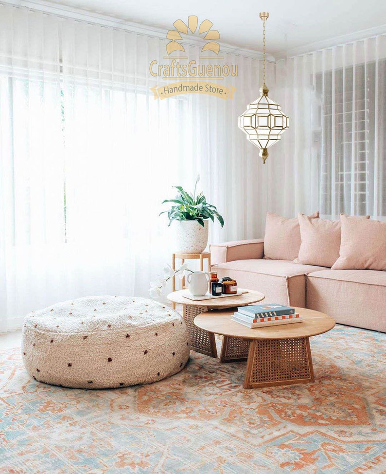 May include: A living room featuring a light pink sofa, a white fluffy ottoman, and two round wooden coffee tables. The room has a large window with white sheer curtains and a white ceiling with a gold geometric pendant light hanging from it. The floor is covered in a colourful rug with a floral pattern.
