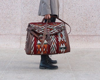 May include: A woman wearing a gray blazer, a red beanie, and black boots is holding a large, colorful, patterned travel bag. The bag has leather accents and straps.