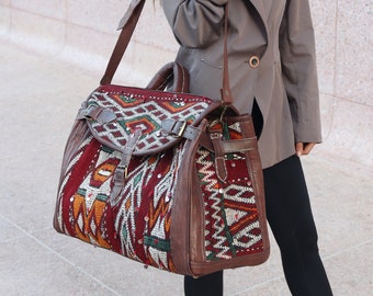 May include: A woman wearing a gray blazer and a red beanie is carrying a large, brown leather bag with a colorful, patterned fabric insert. The bag has multiple buckles and straps.