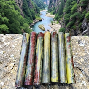 Può includere: Un set di sei pen blank cilindrici in resina caratterizzati da un motivo marmorizzato nelle tonalità verde oliva, giallo e rosso scuro. Le superfici lisce e lucide mostrano variazioni di colore vorticose e pagliuzze metalliche.