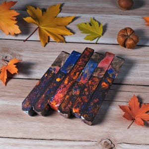 May include: A collection of six rectangular, colorful resin pieces with a metallic edge. The resin features a mix of blues, oranges, and reds, resembling a fiery, abstract design. The pieces are arranged on a wooden surface with autumn leaves.