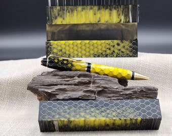May include: A black and yellow pen with a honeycomb pattern. The pen is lying on a piece of wood. There are several blocks of yellow and black resin with a honeycomb pattern in the background.