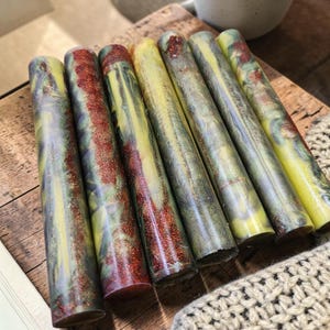Pu&ograve; includere: Sette grezzi cilindrici in resina per penne disposti in fila. Ogni pezzo presenta un motivo marmorizzato con vortici gialli, grigio scuro e verdi, accentuato da striature di glitter scintillante color rame incastonate nel materiale.