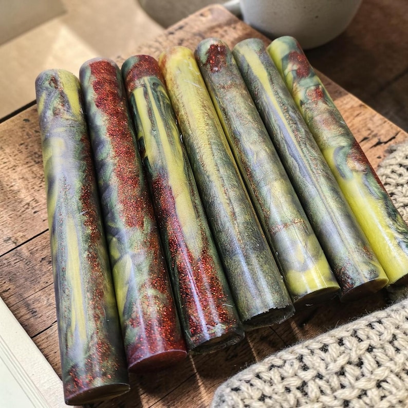 Pu&ograve; includere: Sette grezzi cilindrici in resina per penne disposti in fila. Ogni pezzo presenta un motivo marmorizzato con vortici gialli, grigio scuro e verdi, accentuato da striature di glitter scintillante color rame incastonate nel materiale.