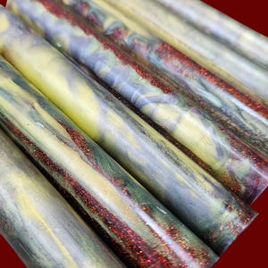 Pu&ograve; includere: Una collezione di cilindri in resina per tornitura caratterizzati da un motivo marmorizzato con vortici gialli, verde scuro e grigi, arricchiti da striature incastonate di glitter rosso scintillante sulle superfici lisce e levigate.