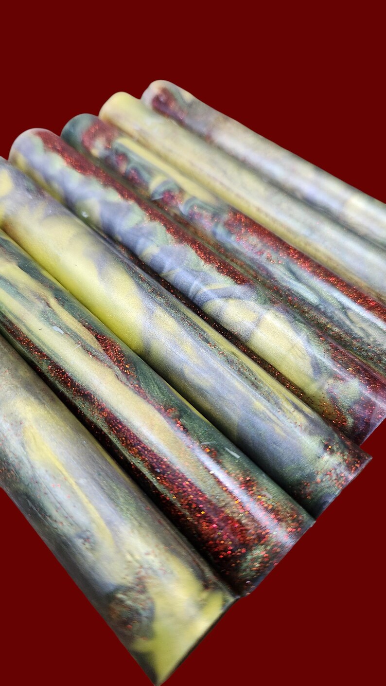 Pu&ograve; includere: Una collezione di cilindri in resina per tornitura caratterizzati da un motivo marmorizzato con vortici gialli, verde scuro e grigi, arricchiti da striature incastonate di glitter rosso scintillante sulle superfici lisce e levigate.