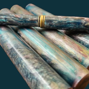 Könnte beinhalten: Eine Sammlung handgefertigter Stifte mit einem einzigartigen Marmordesign. Die Stifte weisen eine Mischung aus Blau-, Grün- und Brauntönen auf, akzentuiert durch goldfarbene Metallkomponenten. Die Stifte sind auf einem dunkel-petrolfarbenen Hintergrund angeordnet, der ihre künstlerische Handwerkskunst zeigt.