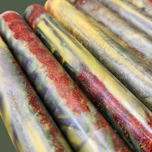 Pu&ograve; includere: Una collezione di cilindri in resina caratterizzati da una miscela marmorizzata di vortici gialli, verdi e viola scuro. Ogni asta &egrave; accentuata da una prominente striscia testurizzata di glitter rosso scintillante che corre lungo la superficie.