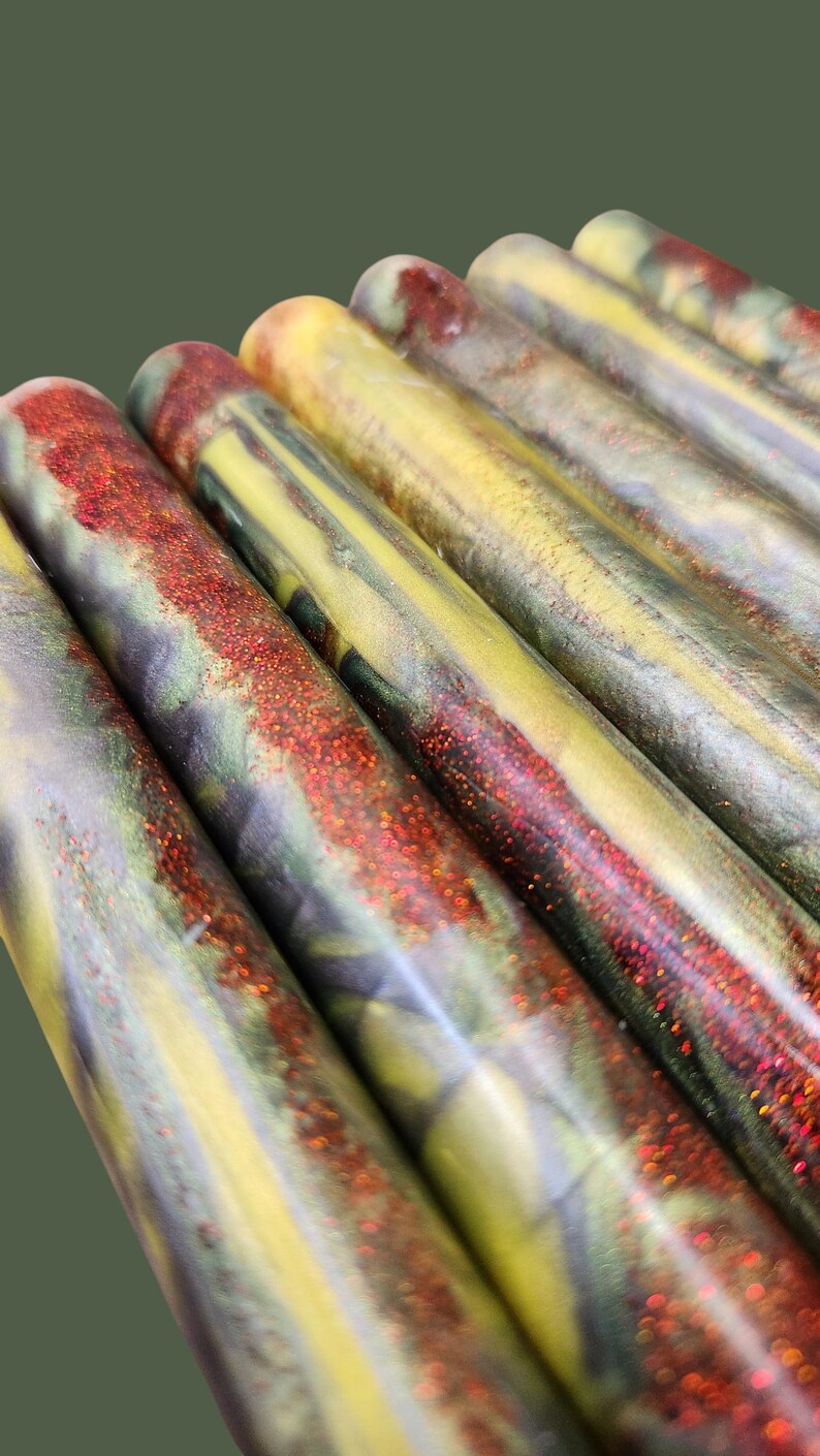 Pu&ograve; includere: Una collezione di cilindri in resina caratterizzati da una miscela marmorizzata di vortici gialli, verdi e viola scuro. Ogni asta &egrave; accentuata da una prominente striscia testurizzata di glitter rosso scintillante che corre lungo la superficie.