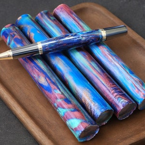 May include: A collection of colorful pens and pen blanks. The pens and blanks feature a swirling pattern of blue, purple, and teal. The pen has a gold-colored tip and accents. The items are arranged on a wooden tray.