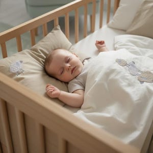 Könnte beinhalten: Ein schlafendes Baby in einem Holzbettchen mit einem beigen Kissen und einer weißen Decke. Das Kissen hat ein Schafdesign. Im Hintergrund ist ein hellblauer Sessel zu sehen.