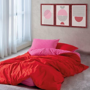 Op de afbeelding: Een rode en roze beddengoedset met een rode dekbedovertrek en twee roze kussenslopen. Het beddengoed ligt op een bed met een grijs tapijt. Er hangen drie ingelijste prints aan de muur achter het bed.