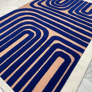 Op de afbeelding: Een blauw en perzikkleurig tapijt met een geometrisch patroon van gebogen lijnen. Het tapijt heeft een witte rand.