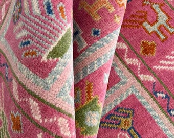 May include: Close-up of a pink rug with a colorful geometric pattern. The rug features a repeating design of stylized animals and geometric shapes in shades of pink, blue, green, and orange.