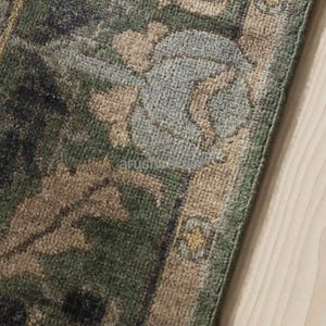May include: Close-up of a section of a green and beige patterned rug. The rug features a floral design with shades of green, beige, and blue. The rug's border is a lighter beige color. The rug is on a light wood surface.