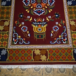 Peut inclure: Tapis rectangulaire marron avec un motif floral et géométrique complexe. Le tapis présente un motif central de vase avec des fleurs, entouré d'une bordure de motifs répétitifs bleus, jaunes et blancs. Le tapis a une frange sur le bord inférieur.