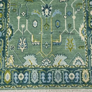 May include: A green rug with a traditional Persian design. The rug has a white fringe around the edges and features a repeating pattern of flowers and leaves.