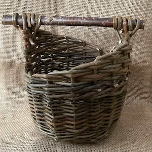 May include: A hand-woven, oval-shaped basket with a wooden handle. The basket is made of natural-coloured woven twigs and branches, with a dark brown handle. The basket is set against a burlap background.