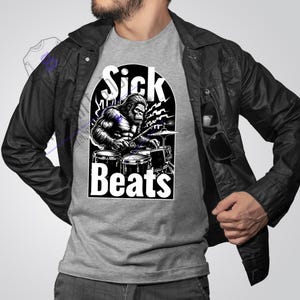 May include: A grey t-shirt featuring a black and white graphic of a gorilla playing drums. The graphic includes the text "Sick Beats".