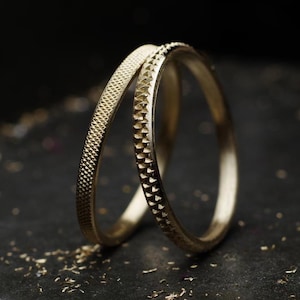 May include: Two gold rings with textured surfaces. One ring has a dotted pattern, and the other has a triangular pattern. The rings are set against a dark background, with small gold flecks scattered around.