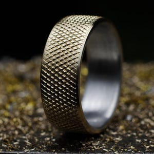Op de afbeelding: Een metalen ring met een getextureerd, kruislings gekarteld patroon op het buitenoppervlak. De ring heeft een breed profiel met een matte goudkleurige afwerking aan de buitenkant en een glad, zilverkleurig binnenoppervlak.