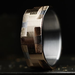 May include: A metallic ring featuring a faceted, geometric outer surface with a polished finish. The band displays a series of interconnected rectangular planes that catch the light, creating a multi-tonal appearance with silver and dark grey hues.
