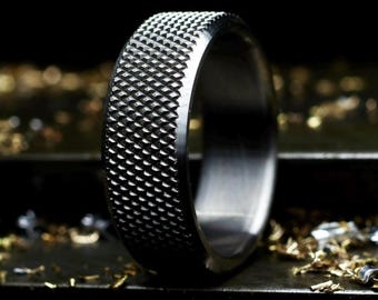 May include: A dark grey metal ring featuring a prominent cross-hatched knurled texture across its outer surface. The band has a wide, flat profile with a smooth, polished interior finish, showcasing a distinct industrial aesthetic and metallic sheen.