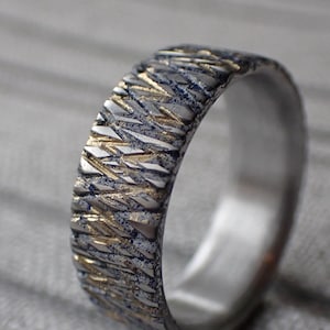 May include: A silver and gold wedding band with a textured, geometric pattern. The band is made of two metals, with the gold accents creating a contrasting design.