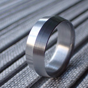 May include: A silver ring with a beveled edge and a brushed finish. The ring is lying on a textured surface.