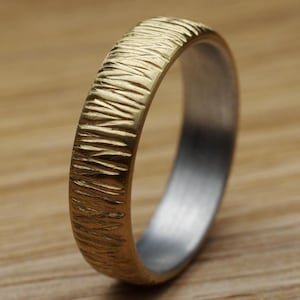 Könnte beinhalten: Ein goldfarbener Ring mit strukturierter Oberfläche. Der Ring hat ein goldenes Band mit strukturiertem Muster und einen silberfarbenen Innenraum. Der Ring ist vor einem Holzgrund platziert.