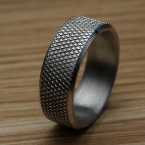 May include: A close-up shot of a silver ring with a textured, diamond-patterned surface. The ring is made of a shiny, metallic material and sits on a wooden surface. The ring's design is simple yet detailed.